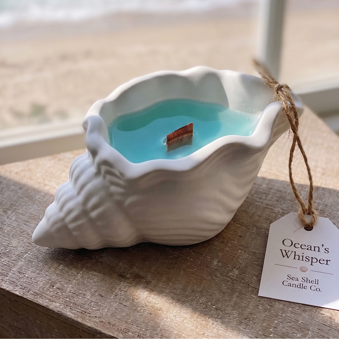 A seashell-shaped candle with light blue wax sits on a wooden surface in front of a window with a beach and ocean view in the background. A paper tag on twine reads &