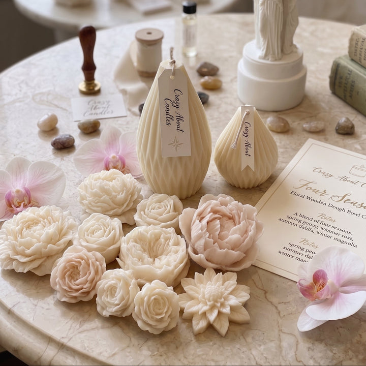 A styled tabletop display of cream and blush wax melts shaped like flowers, alongside a white statue candle holder, beeswax candles, a wax seal stamp, and printed cards on a marble surface.