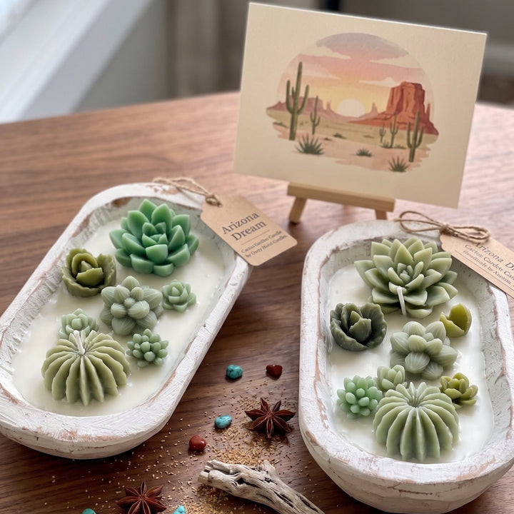 Two white oval dish-shaped soaps decorated with various green succulent and cactus designs, displayed on a wooden table alongside a desert landscape art print on a small easel.