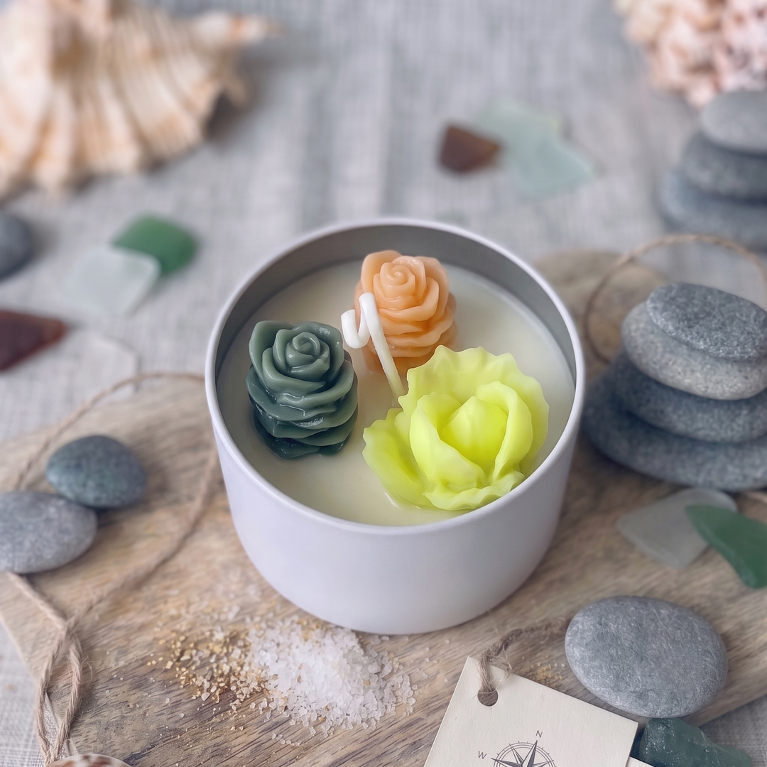 A white ceramic candle tin filled with wax and decorated with three sculpted flower wax ornaments in green, peach, and pale yellow, styled on a wooden board with sea glass and stones.