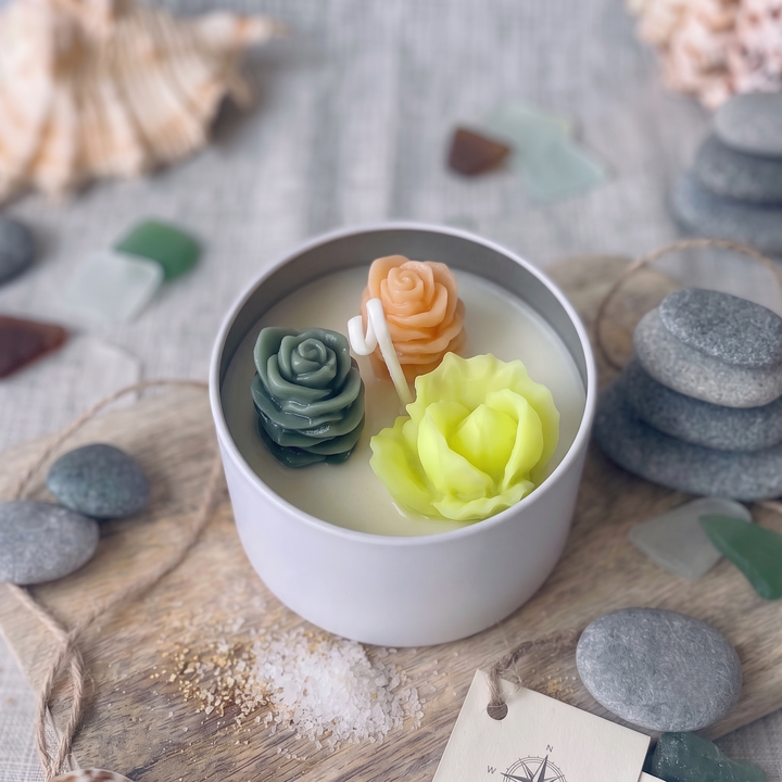 A white ceramic candle tin filled with wax and decorated with three sculpted flower wax ornaments in green, peach, and pale yellow, styled on a wooden board with sea glass and stones.