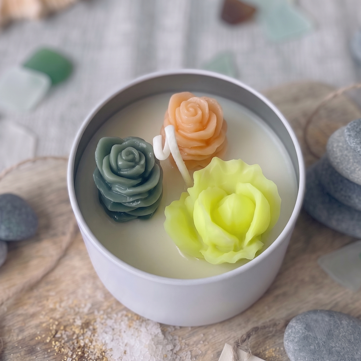 A white candle tin filled with creamy wax and three sculpted rose-shaped wax candles in sage green, pale peach, and chartreuse yellow, styled on a wooden surface with smooth stones.