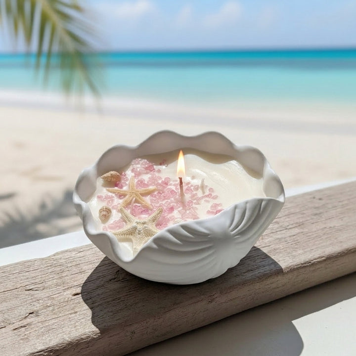 A seashell-shaped candle with pink starfish and sea urchin accents, lit with a small flame, set on a wooden railing with a blurred beach and ocean background.