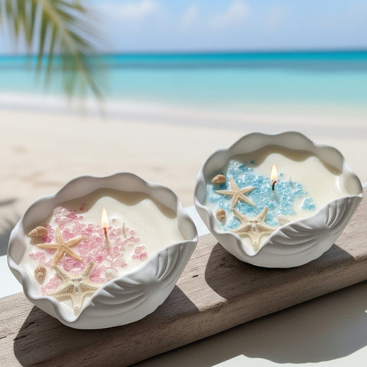 Two seashell-shaped candles lit on a wooden board with a tropical beach background. One candle has pink starfish and beads, and the other has blue starfish and beads.