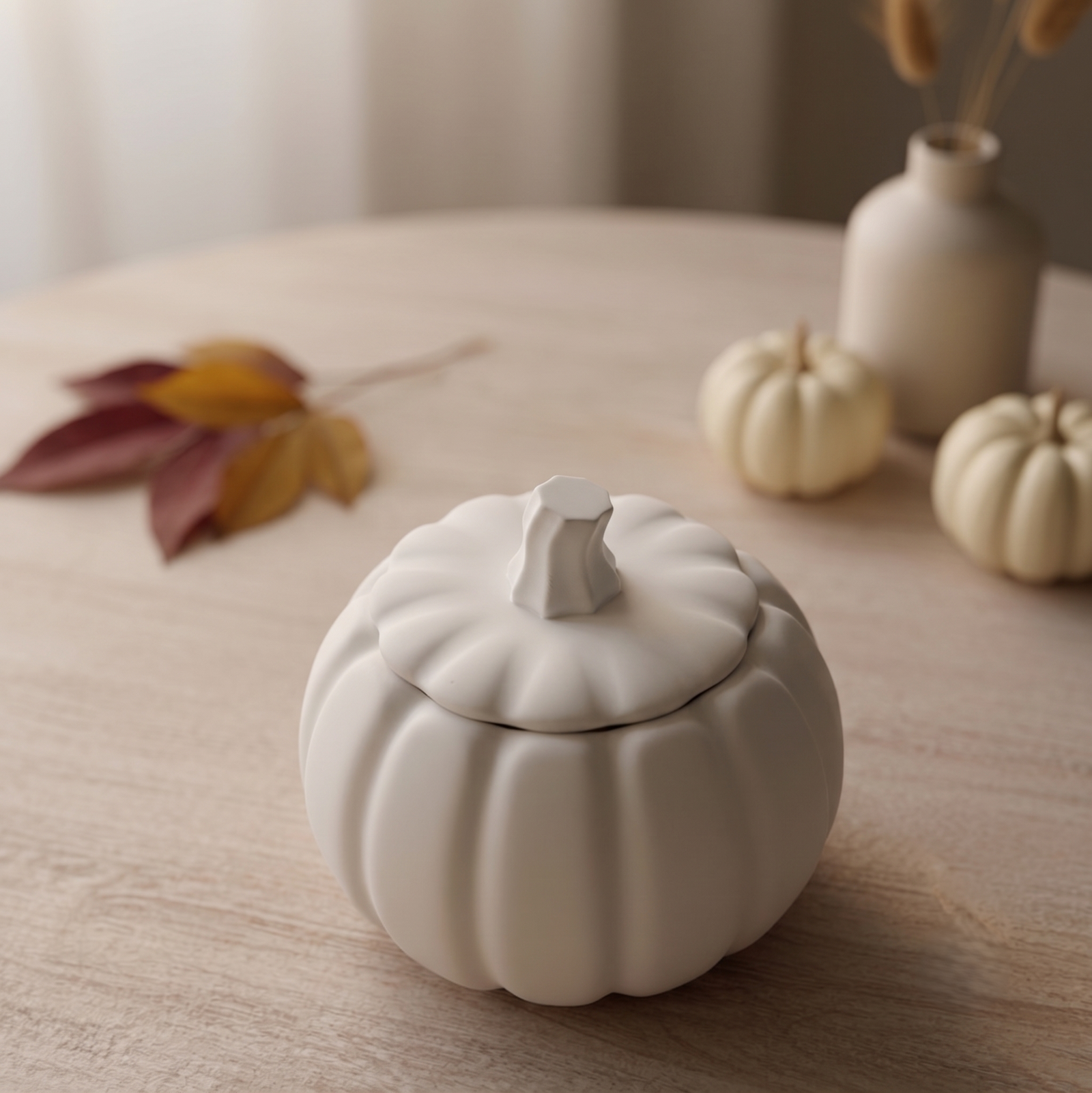 A white ceramic pumpkin-shaped lidded container sits on a light wood table, styled with autumn leaves and small decorative pumpkins in the background.