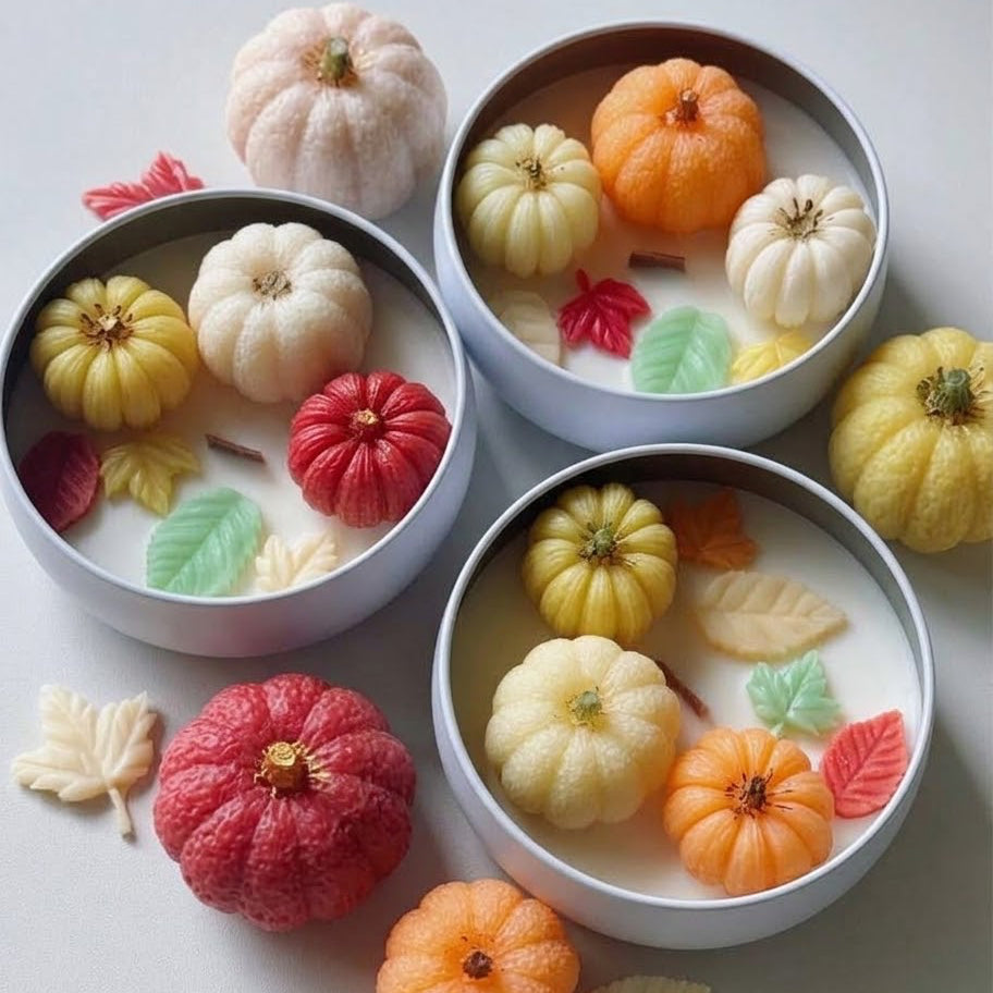 Three round metal tins filled with creamy white wax and decorated with small sculpted pumpkins and fall leaves in orange, yellow, red, and cream, arranged on a light neutral background with additional decorative pumpkins and leaves around them.
