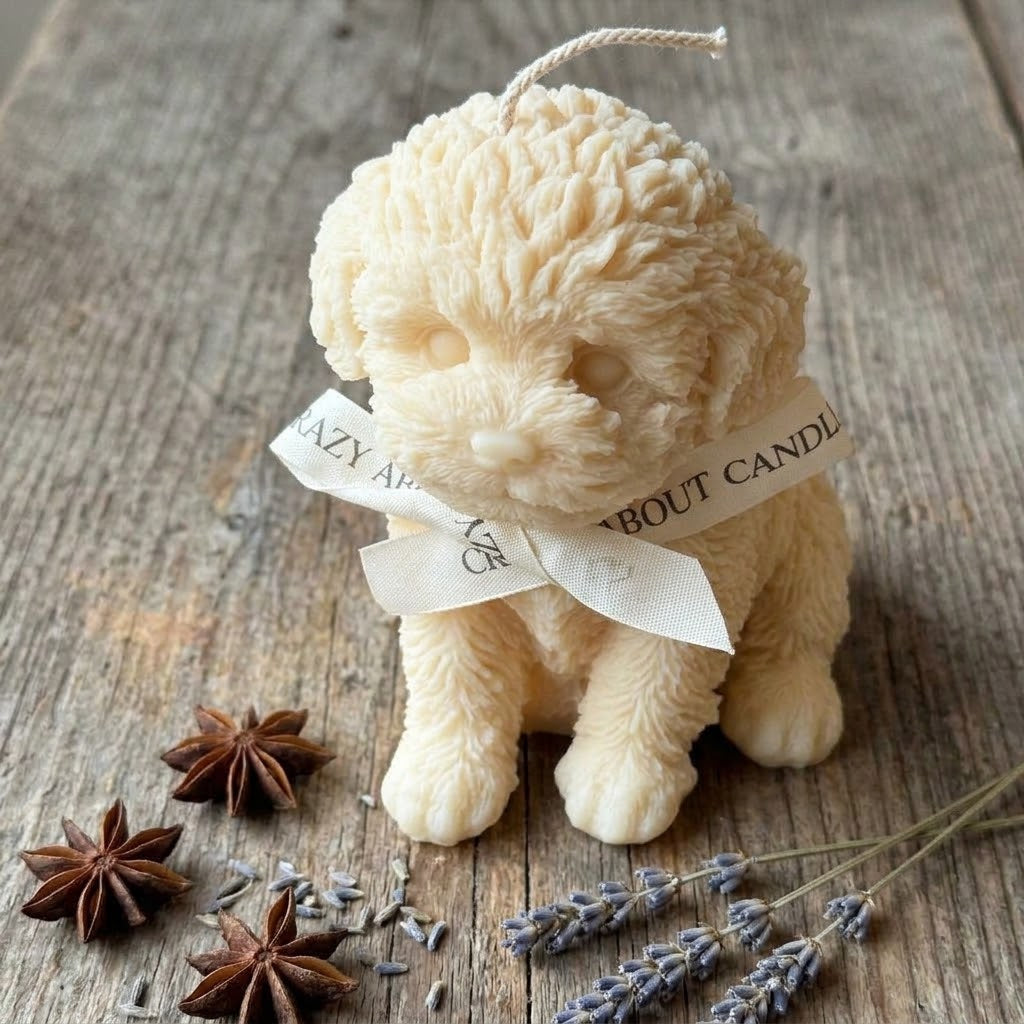 A sculpted cream-colored dog-shaped candle with a hanging loop and a white ribbon bow tied around its neck, placed on a rustic wooden surface with star anise and lavender sprigs nearby. The ribbon reads &