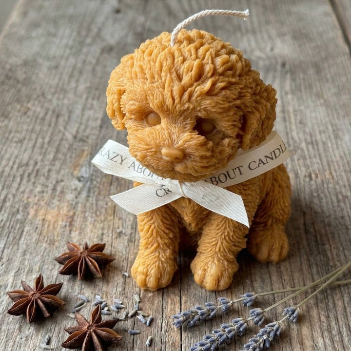 A sculpted dog-shaped candle in warm tan/golden brown sits on a rustic wooden surface, tied with a cream ribbon that reads &