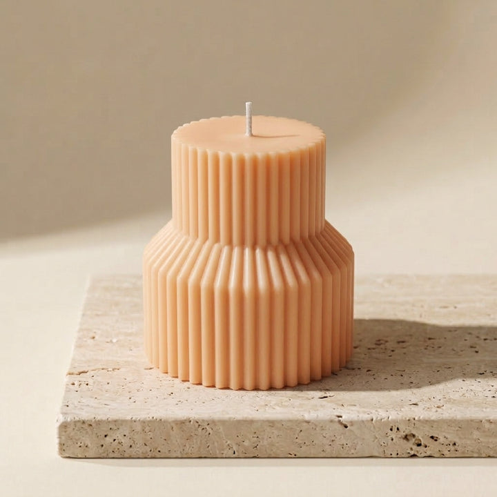 A two-tiered ribbed candle in a warm peach-beige color, displayed on a textured stone slab against a neutral beige background.