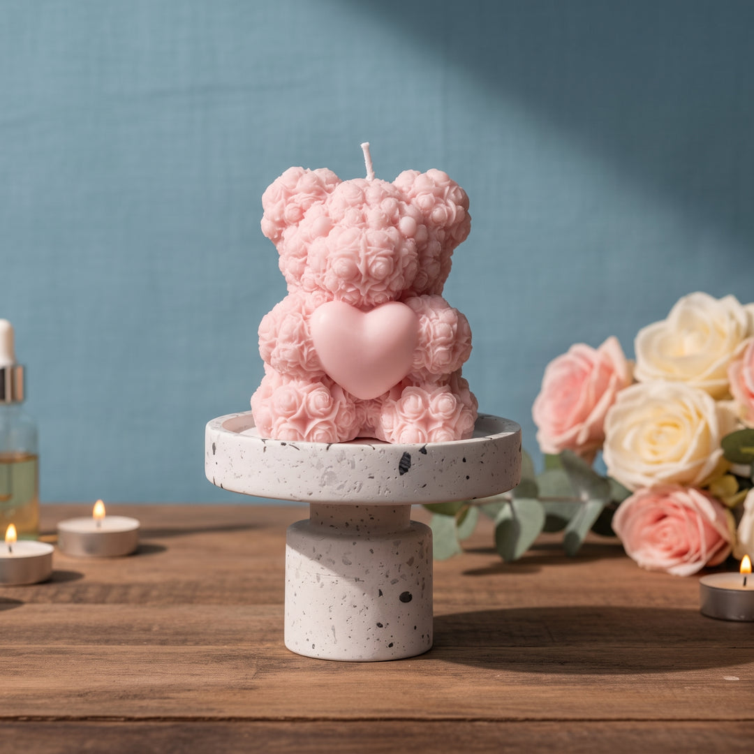 A pink teddy bear candle with a heart-shaped center sits on a white speckled pedestal. The scene is styled on a wooden table with small lit candles, a bottle, and pink and white roses against a blue wall.