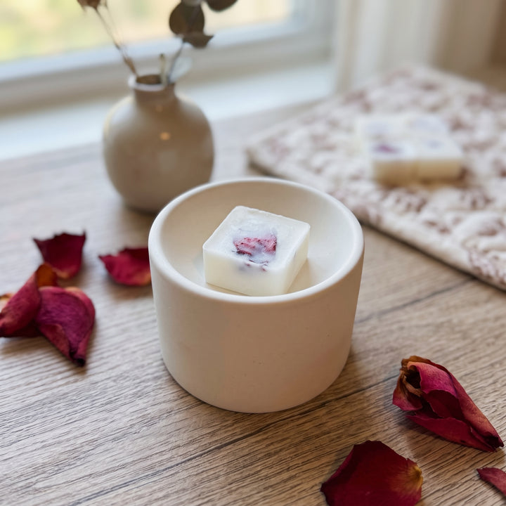 A small white square soap bar with a pinkish-red flower embedded in it sits in a matte cream ceramic dish on a wooden table, with dried rose petals scattered around and a softly blurred background scene.