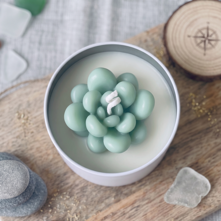 A round white candle tin filled with pale mint-green wax, featuring a sculptural rosette-shaped wax top in matching mint green, styled on a wooden surface with stones and a wood slice.