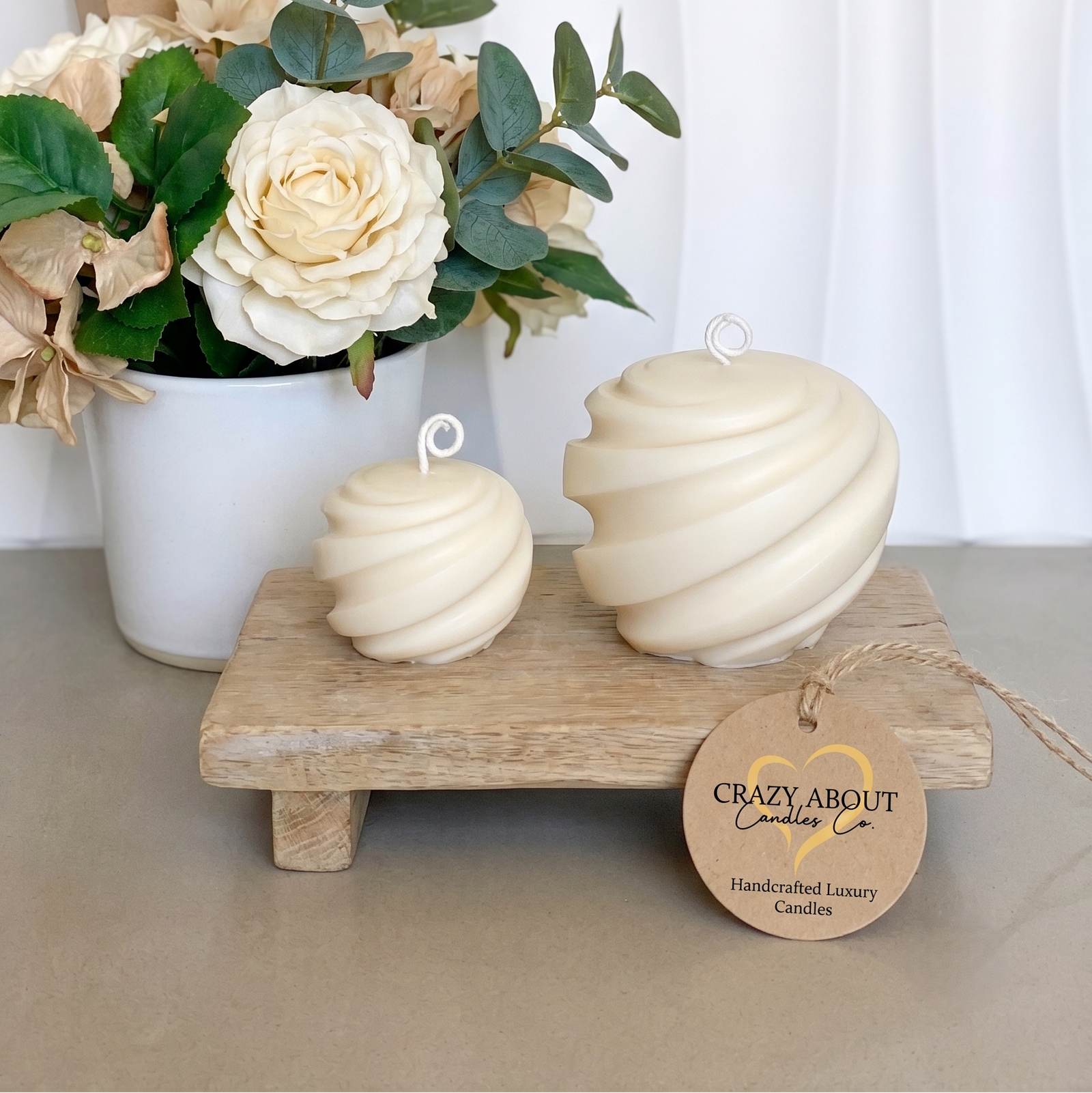 Two cream-colored spiral twist candles displayed on a small wooden stand, with a matching branded hang tag. A white flower arrangement in a vase and a kraft gift box are styled in the background against a white curtain backdrop.