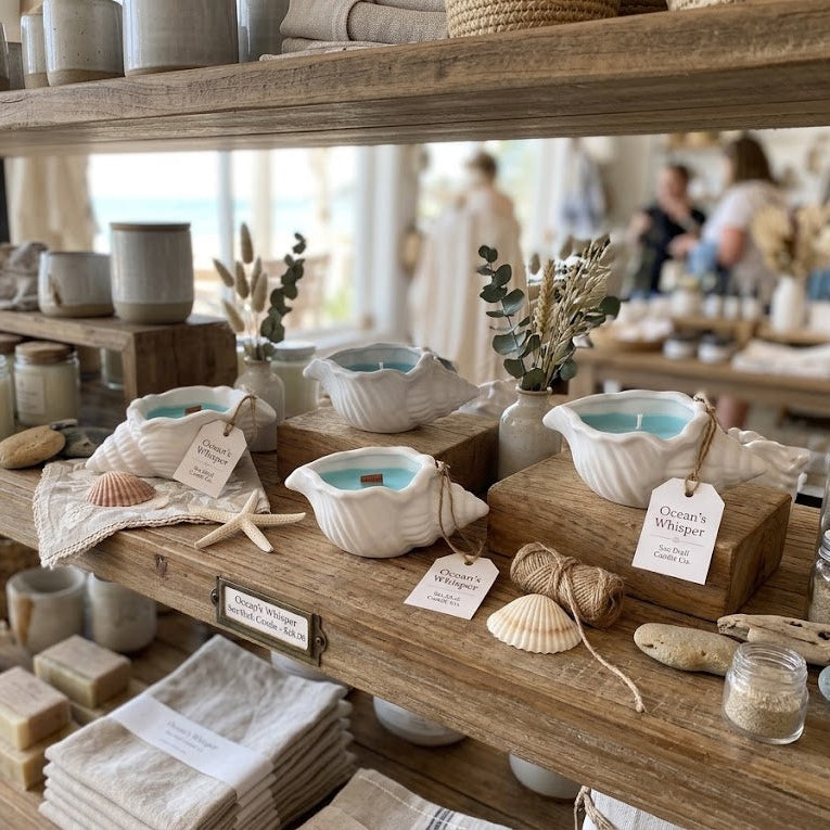 A styled retail display on rustic wooden shelves featuring various home and bath products, including ceramic bowls with light blue interiors, candles, soap bars, folded towels, and small decorative seashells and dried grass in a softly blurred boutique setting.