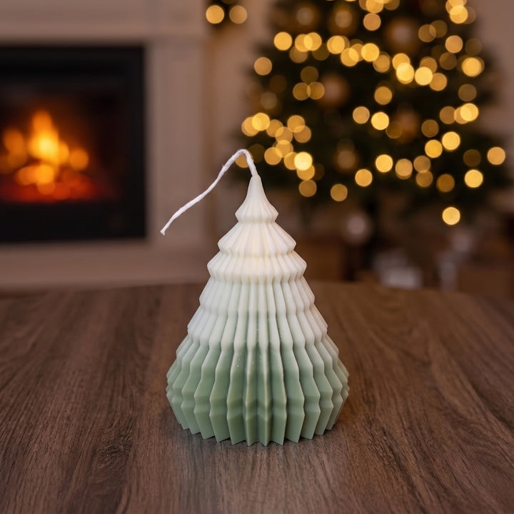 A Christmas tree-shaped candle with a white top fading to a light sage green base, styled on a wooden table with a blurred fireplace and festive lights in the background.
