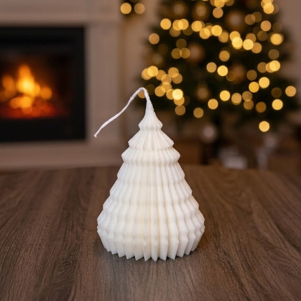 A white Christmas tree-shaped candle with a textured, pleated design sits on a wooden table in a cozy living room setting with a blurred fireplace and a lit Christmas tree in the background.