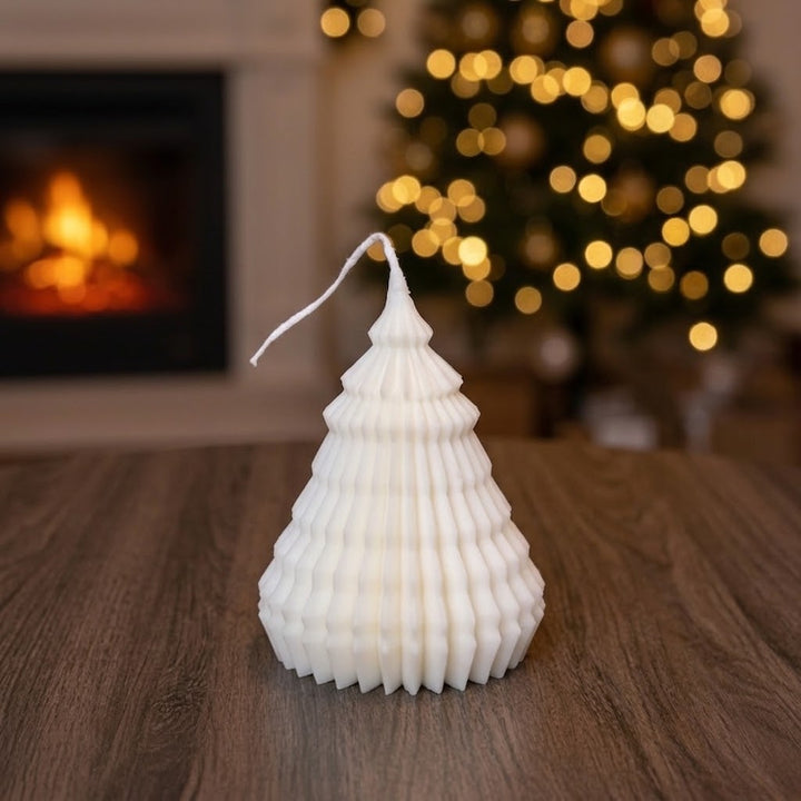 A white Christmas tree-shaped candle with a textured, pleated design sits on a wooden table in a cozy living room setting with a blurred fireplace and a lit Christmas tree in the background.