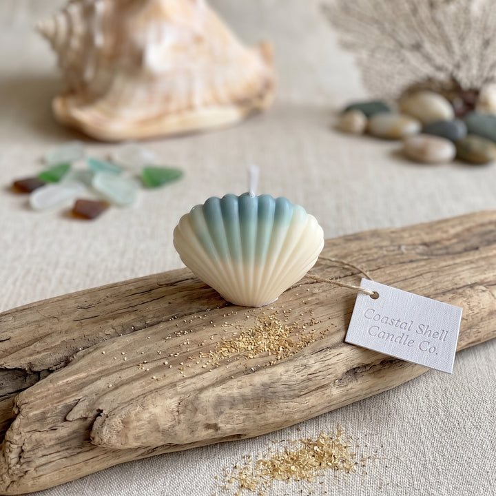 A seashell-shaped candle with a blue-to-cream gradient sits on a piece of driftwood with a paper tag reading &