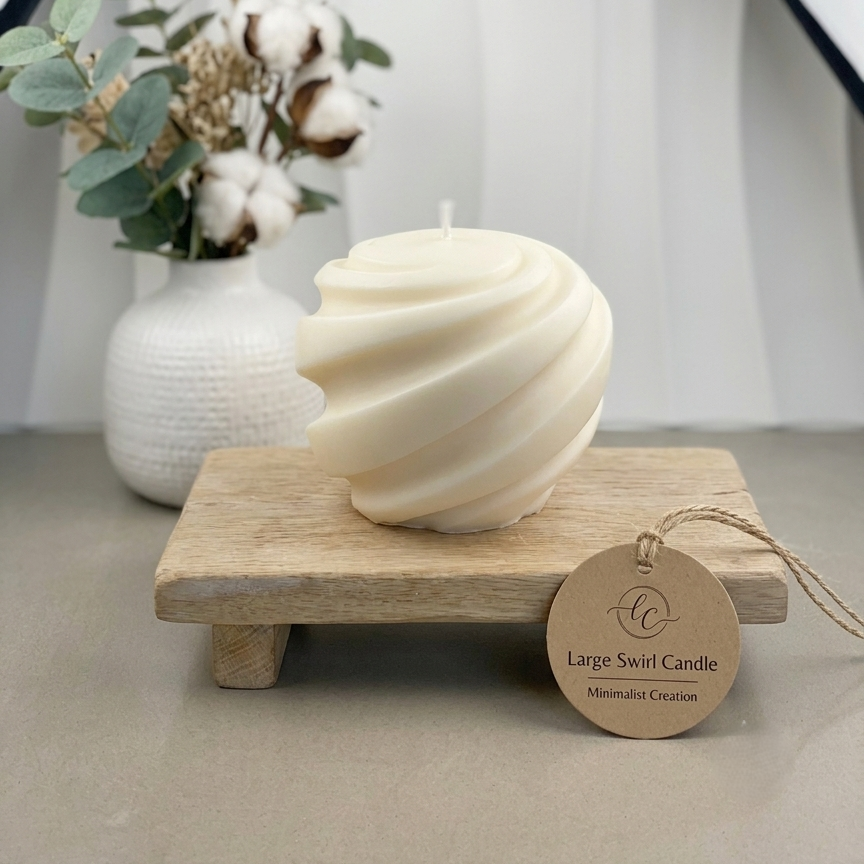 A large cream-colored spiral swirled candle displayed on a small wooden stand, with a round kraft paper tag attached. The setup is styled with a white textured vase containing cotton bolls and green leaves in the background against a draped white fabric backdrop.