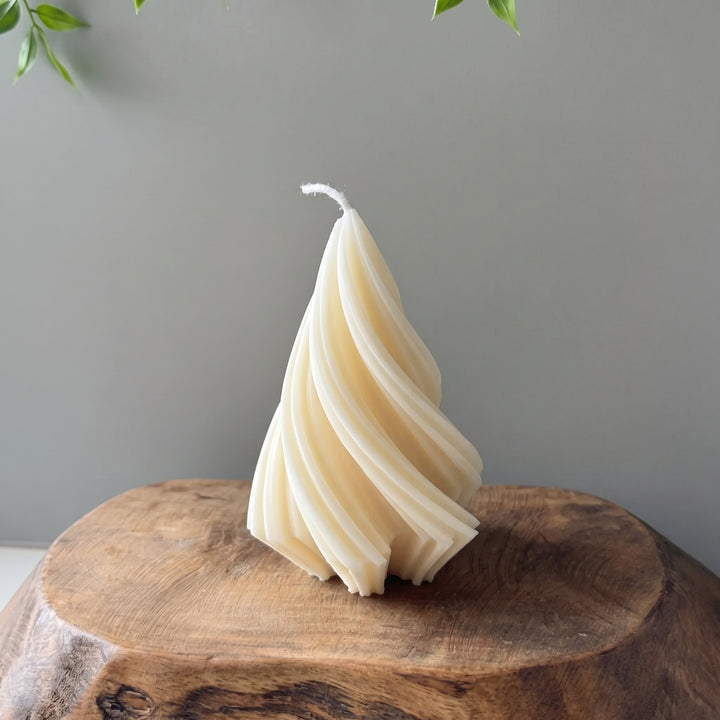 A sculptural spiral-shaped candle in a warm cream or ivory tone, displayed on a rustic wooden slab against a plain gray wall.