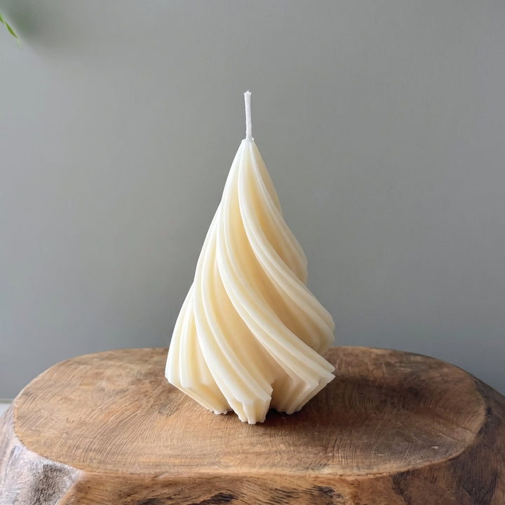 A sculptural spiral candle made of layered cream-colored wax stands on a rustic wooden slice against a plain gray wall.