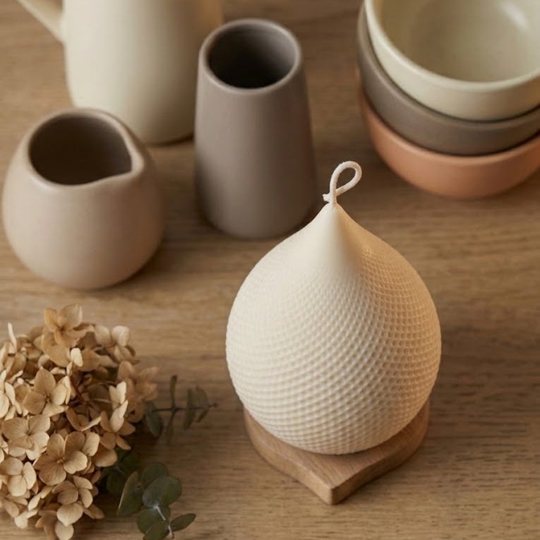 A textured cream-colored ceramic onion-shaped container with a hanging loop, displayed on a wooden coaster on a wooden tabletop. In the background, a matching cream pitcher, a gray vase, and stacked bowls in beige, brown, and terracotta are arranged with dried flowers.