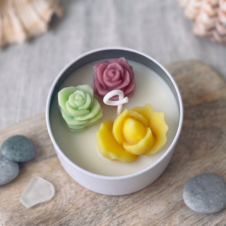 A white candle in a round white tin with three sculpted flower wax ornaments in green, pink, and yellow, styled on a wooden surface with stones and shells.
