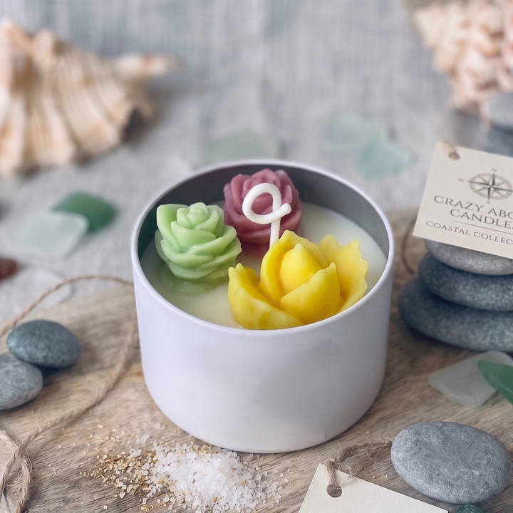 A white ceramic candle vessel filled with wax and topped with three flower-shaped candles in green, pink, and yellow, styled on a wooden surface with seashells, rocks, and a tag reading &