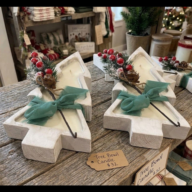 Two white-painted wooden Christmas tree-shaped candle holders on a rustic wooden table, each filled with a white candle and decorated with a teal bow, green pine sprigs, and red berries. A small tag reads 'Tree Bowl Candle $32'.
