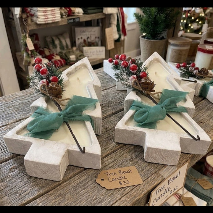 Two white-painted wooden Christmas tree-shaped candle holders on a rustic wooden table, each filled with a white candle and decorated with a teal bow, green pine sprigs, and red berries. A small tag reads &