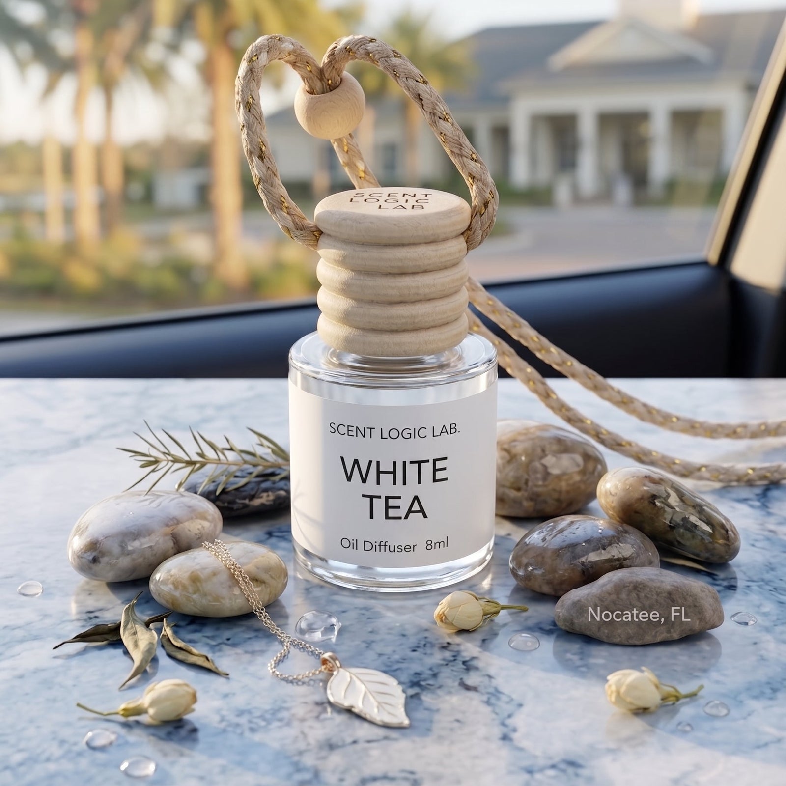 A Scent Logic Lab White Tea oil diffuser bottle with a wooden diffuser top and rope loop, styled on a marble surface with stones, dried leaves, and white tea buds in front of a car window with a blurred outdoor background.
