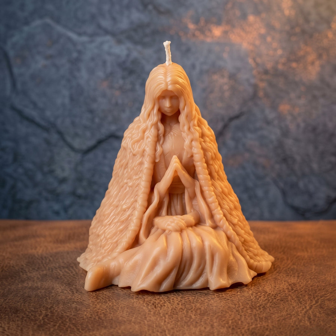 A beige wax candle sculpted as a seated female figure with long flowing hair and a draped cloak, photographed on a textured brown surface against a mottled gray-blue background.