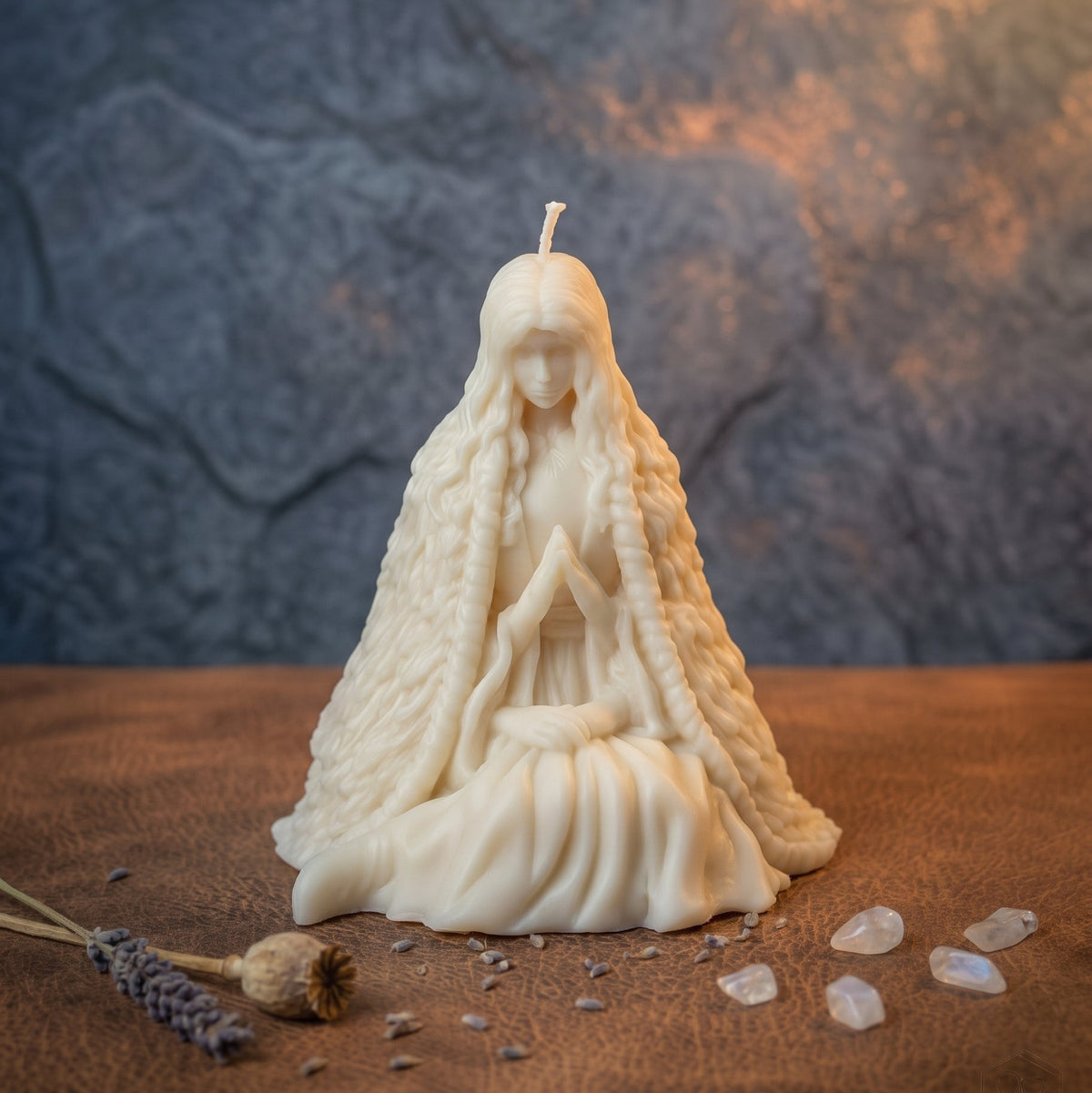 An off-white wax candle sculpted as a seated female figure with long braided hair and a draped robe, placed on a brown textured surface with small crystals and dried lavender around it. The background is a mottled gray wall with warm light reflections.