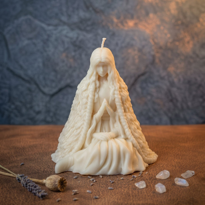 An off-white wax candle sculpted as a seated female figure with long braided hair and a draped robe, placed on a brown textured surface with small crystals and dried lavender around it. The background is a mottled gray wall with warm light reflections.