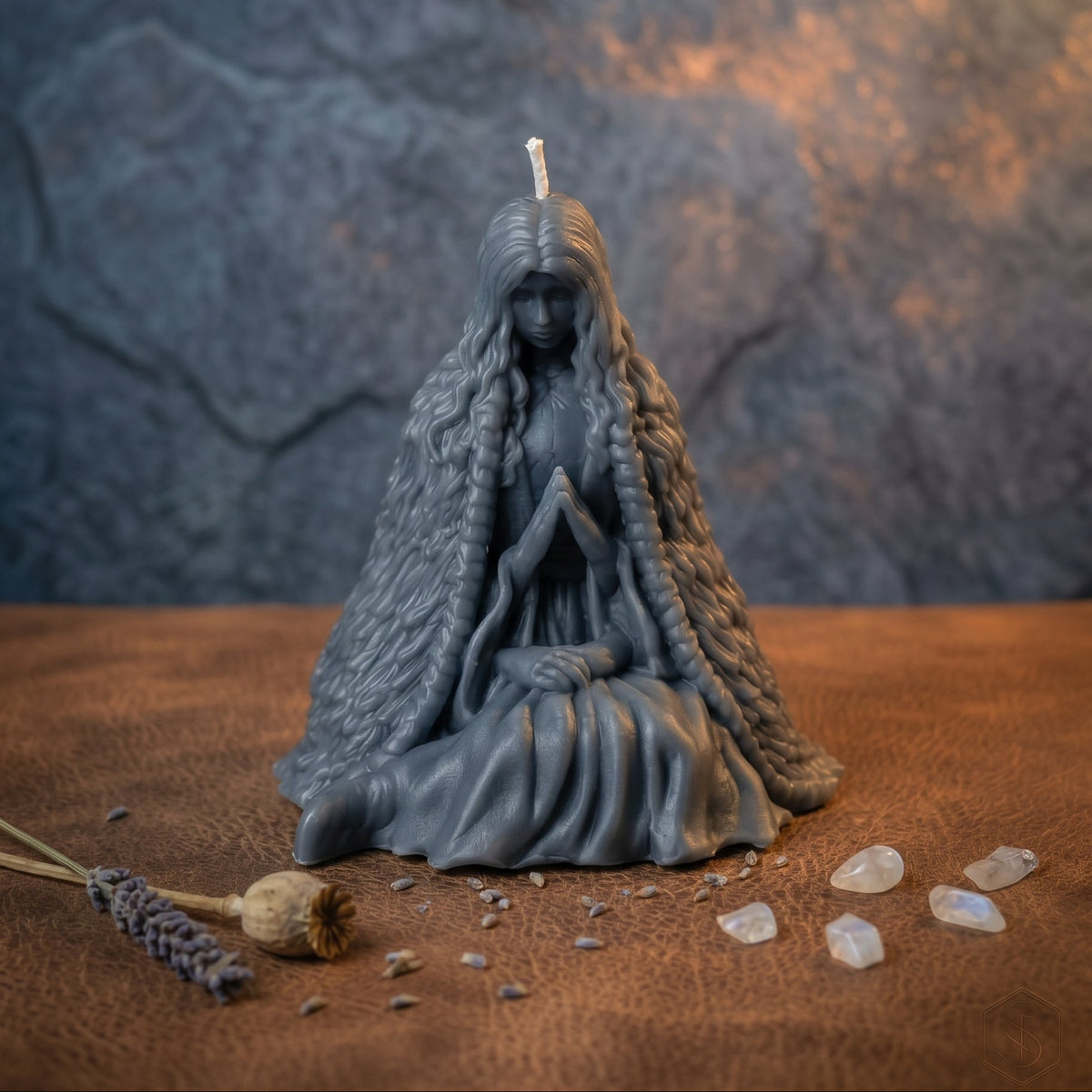 A dark gray figurine candle depicting a kneeling woman with long braided hair and a textured cloak, set on a brown surface with crystals and dried lavender against a textured gray and orange background.