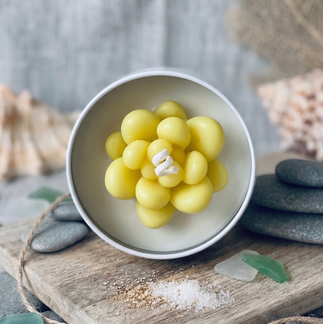 A round white candle tin filled with bright yellow wax, styled on a rustic wooden board with sea glass, rocks, and shells against a gray background.