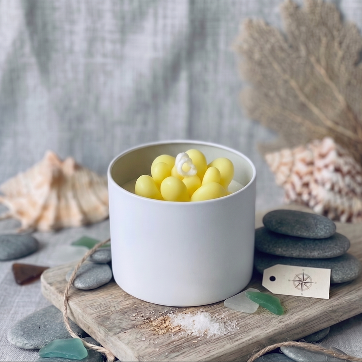 A white ceramic candle holder or candle container filled with pale yellow seashell-shaped wax or beads, styled on a wooden board with sea glass, stones, and dried coral against a soft gray fabric backdrop.