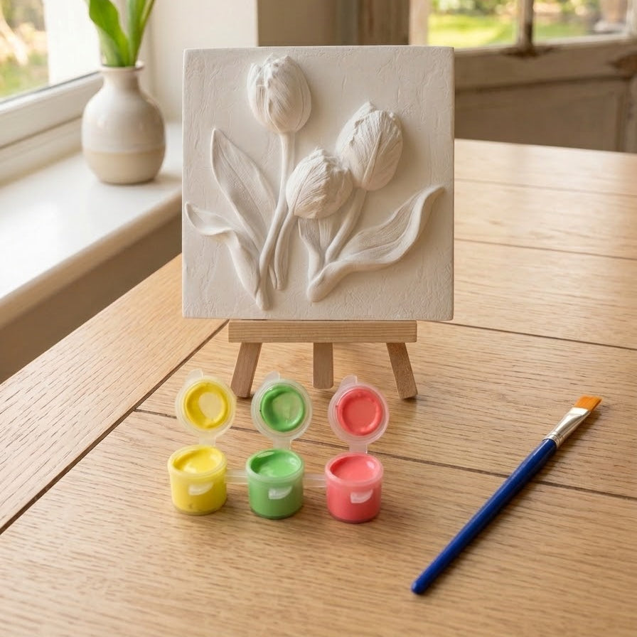 A white painted relief plaque of three tulips on a small wooden easel, set on a wooden table with paint pots, a paintbrush, and a yellow tulip in the background near a window.