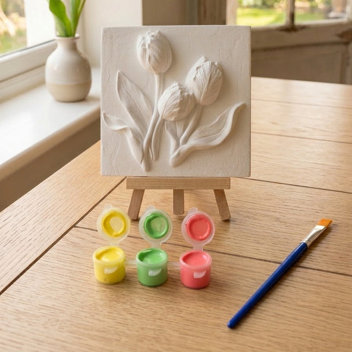 A white painted relief plaque of three tulips on a small wooden easel, set on a wooden table with paint pots, a paintbrush, and a yellow tulip in the background near a window.